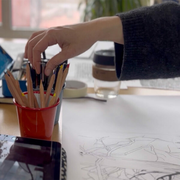 A person reaches for colored pencils on a desk with drawing materials, sketches, a reusable cup, and other art supplies.