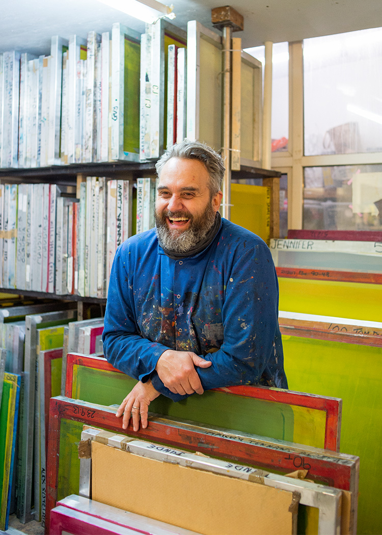 A bearded man in a paint-splattered blue shirt stands smiling among stacked, colorful screen printing frames in a workshop.