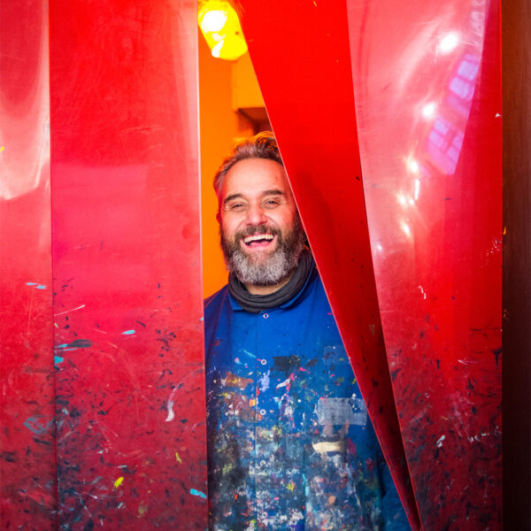A man with a beard and blue paint-splattered shirt stands smiling between two large red curtains in a brightly lit room.