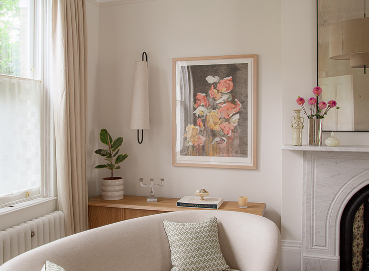 A cozy living room corner with a curved sofa, a wooden console table, a potted plant, Print Club London wall art of flowers, and a marble fireplace decorated with vases and flowers.