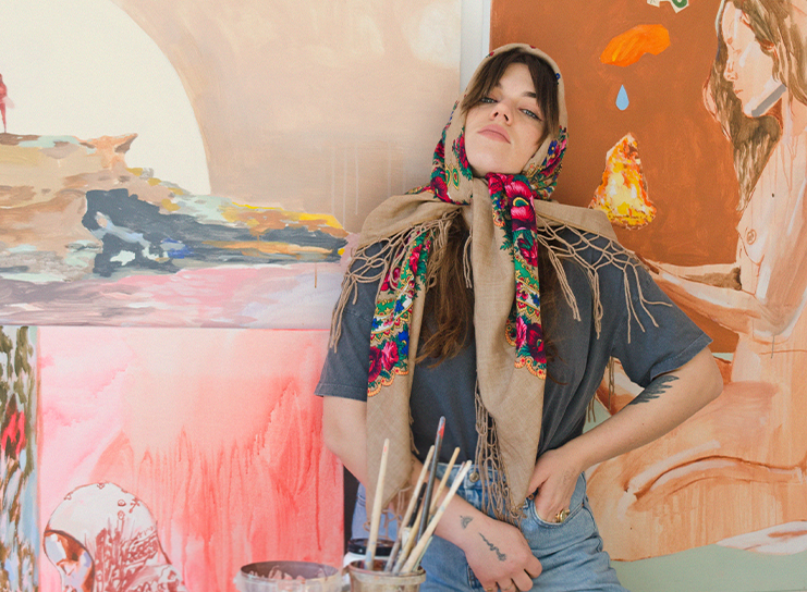 A woman with a floral scarf over her head stands in front of colorful paintings, with paintbrushes and art supplies on a table in the foreground, capturing the creative spirit of Print Club London.