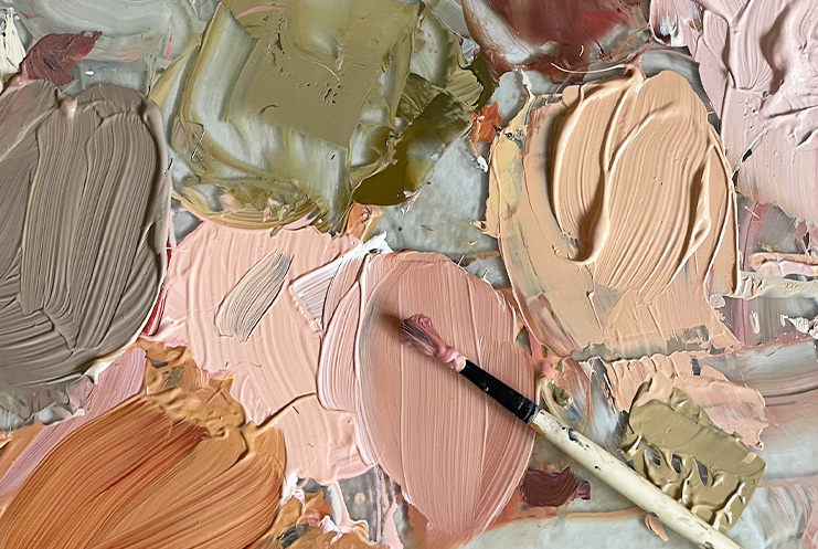 A paintbrush rests on a palette covered in various shades of beige, pink, brown, and olive-colored oil paints—reminiscent of the creative spirit found at Print Club London.