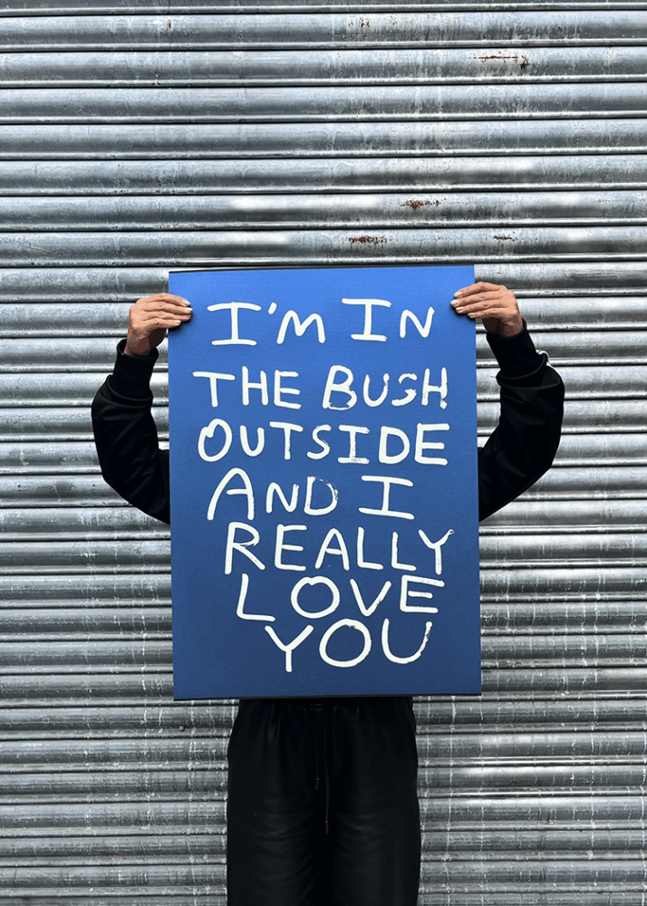 A person in black clothing holds a blue sign with white text reading, "I'M IN THE BUSH OUTSIDE AND I REALLY LOVE YOU," standing in front of a corrugated metal wall.