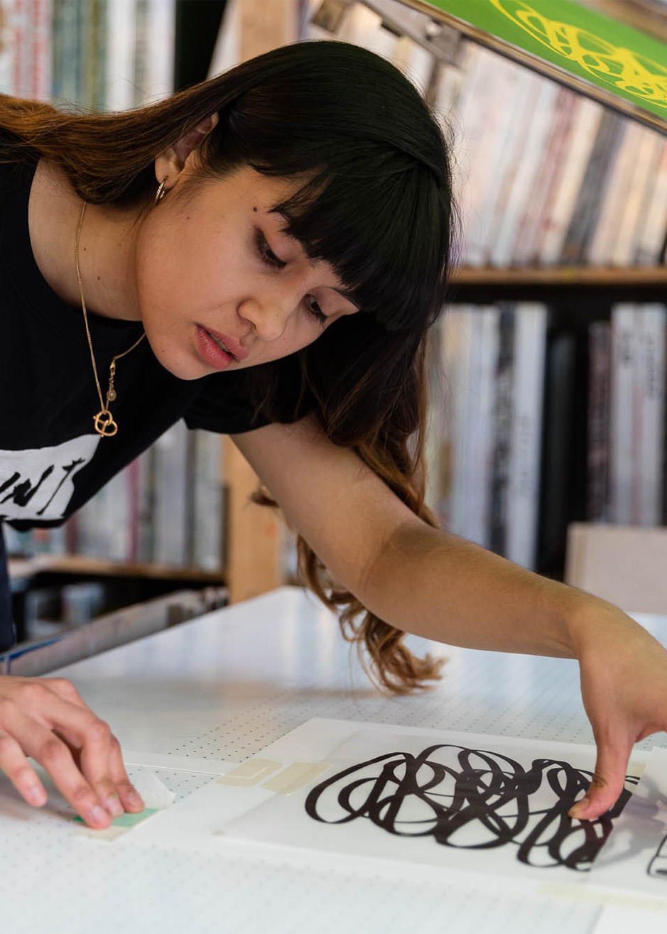 A woman aligns a black abstract sheet on a worktable, possibly prepping for the "Inky Evenings - 2hr beginners workshop." Shelves with materials are visible in the background.