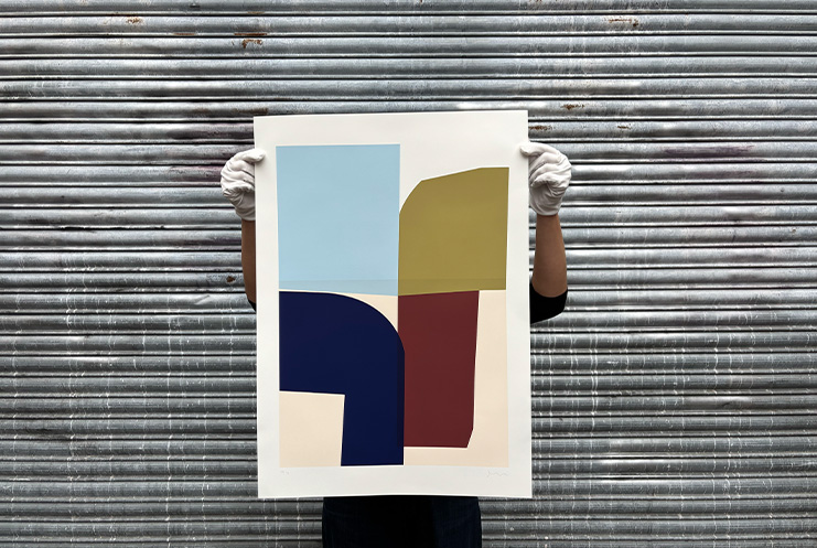 A person wearing white gloves holds up an abstract geometric artwork from Print Club London in front of a corrugated metal wall.
