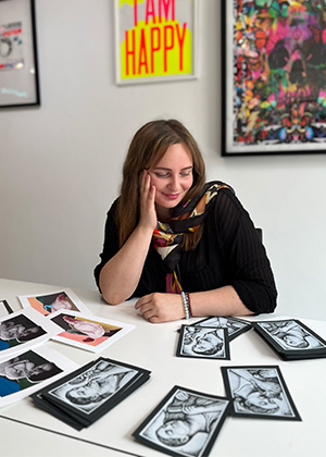 A woman sits at a table looking at several portrait sketches and printed photographs, with colorful artwork hanging on the wall behind her.