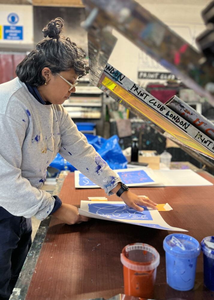 A person in paint-splattered clothes works with a stencil at a table, surrounded by art supplies and prints. A sign reads "Paint Club London.
