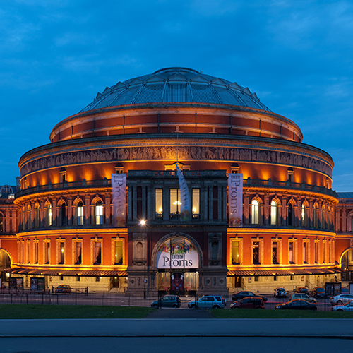 The Royal Albert Hall illuminated at dusk, with charity and community banners for the Proms hanging on the facade and cars parked in front.