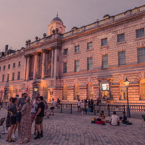 People gather and relax on the cobblestone courtyard in front of a large, historic building at dusk, drawn together by a sense of charity and community.