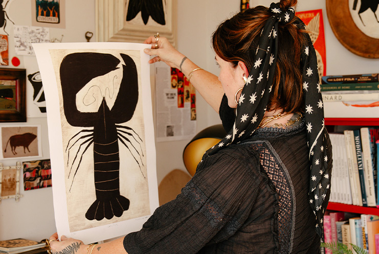 A person with a star-patterned scarf examines a black and white abstract lobster artwork by Print Club London in a room decorated with books and art.