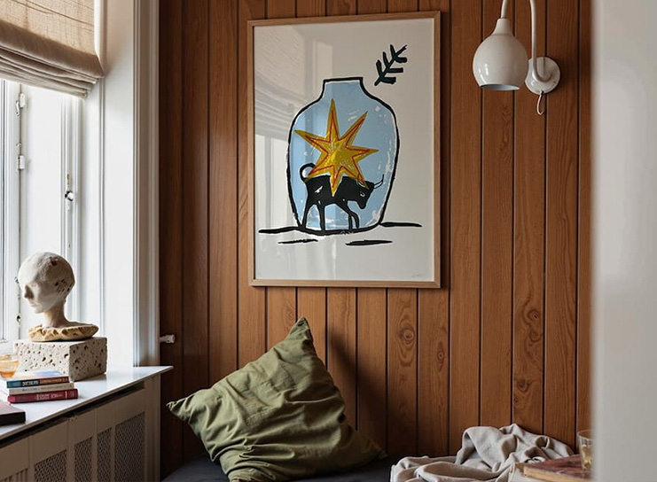 A cozy corner with wood-paneled walls, a limited edition art print of a vase with a camel and star, a sculpture bust, books, and a green cushion on a bench.