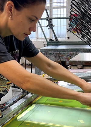 A person uses a squeegee to push ink through a screen in a printmaking studio, with drying racks and equipment visible in the background.