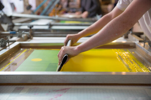 A person uses a squeegee to spread yellow ink across a screen in a screen printing process, showing why real people matter in creating unique, handcrafted prints.
