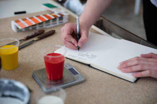 A person writes in a notebook beside cups of paint, a scale, a color palette, and art tools on a table—showing why real people matter in the creative process.