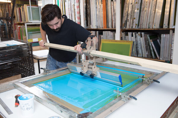 A man operates a screen printing press, spreading blue ink over a large screen in a workshop filled with screens and supplies, showing why real people matter in bringing unique creations to life.