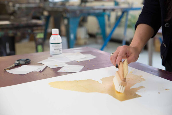 A person uses a wide brush to apply a yellowish liquid to white paper on a table, surrounded by scissors, paper scraps, and adhesive—showcasing why real people matter in the creative process.