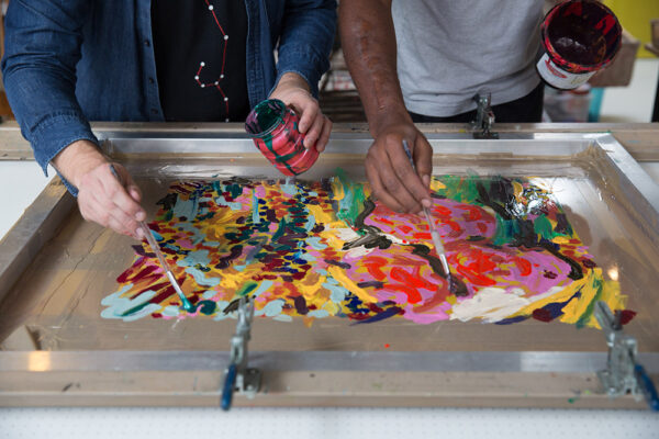 Two people use brushes to apply colorful paint onto a screen printing frame, showing why real people matter in collaborative art projects.