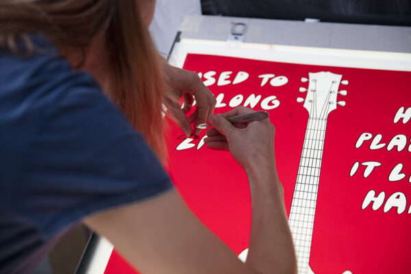 A person uses a tool to arrange white letters on a red sign featuring a guitar illustration and partially visible text, highlighting why real people matter in creating authentic, engaging displays.