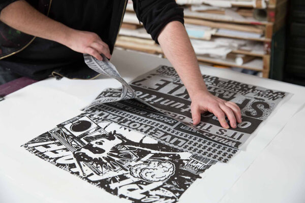 A person lifts a printed sheet with bold black and white graphic text and images from a flat surface in a printmaking studio, capturing the essence of why real people matter in creative expression.