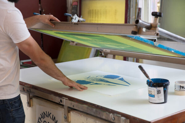 A person uses a screen printing press to create a graphic print on paper, showing why real people matter—crafting unique art by hand, with ink and tools spread out on the table nearby.