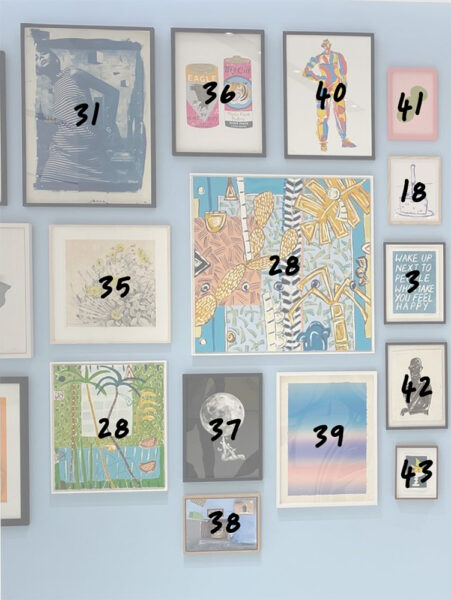 A gallery wall displays various framed artworks from The Bishopsgate Collection art, with numbers written in black marker on the glass of each frame. The paintings and prints vary in style, size, and subject.