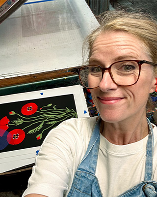 A person wearing glasses and an apron smiles at the camera, standing next to a colorful print of red poppies on a black background.