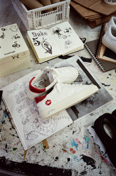 A white trainer with a red lipstick mark and signature sits on top of tattoo sketchbooks and drawings on a cluttered workspace.