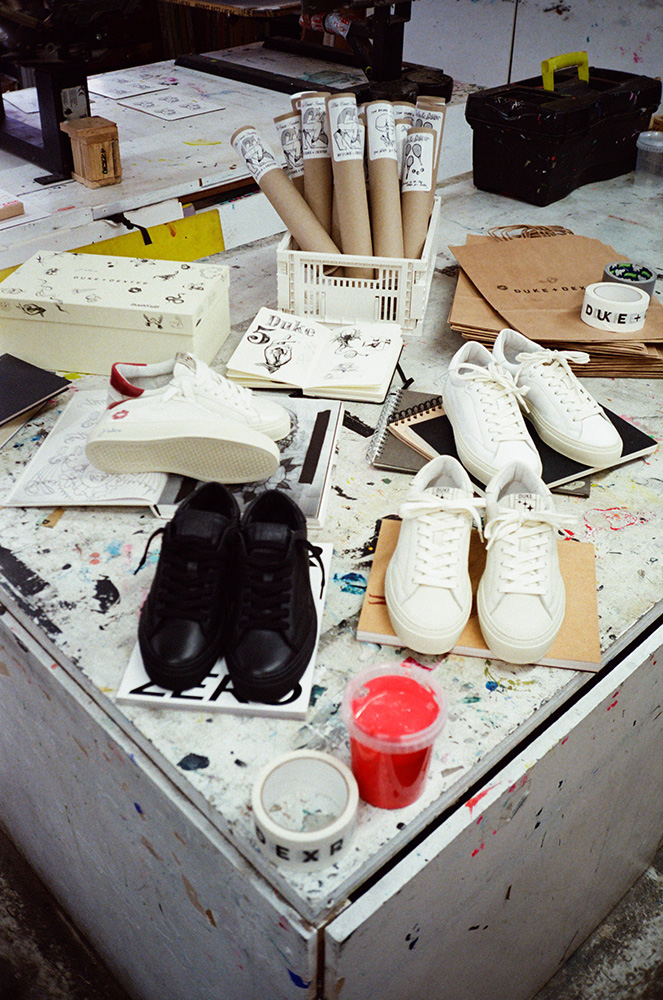 A cluttered worktable with black and white trainers, sketches, cardboard tubes, envelopes, notebooks, a cup of red paint, and other art supplies scattered across the surface.