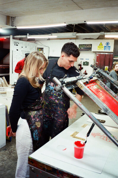Two people wearing aprons stand by a screen printing press in an art studio, looking at prints on the table. A red cup and printed materials are visible.