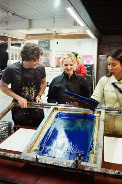 Three people stand around a large screen printing frame with blue ink, working together in an art studio.