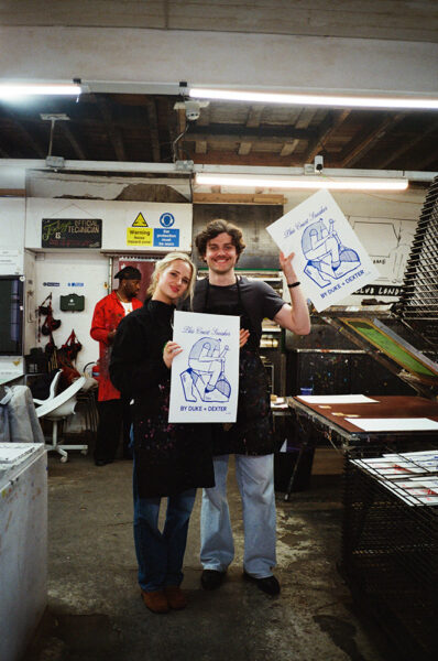 Two people wearing aprons stand in an art studio, smiling and holding up prints with "BY DUKE & DEXTER" text. Art supplies and drying racks are visible in the background.