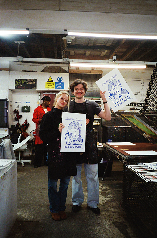 Two people wearing aprons stand in an art studio, smiling and holding up prints with "BY DUKE & DEXTER" text. Art supplies and drying racks are visible in the background.