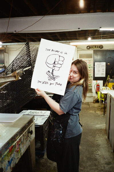 A person in an art studio holds up a poster featuring a drawing of a fist and the text "The Answer is in The Steps you Take." Drying racks and studio equipment are visible.