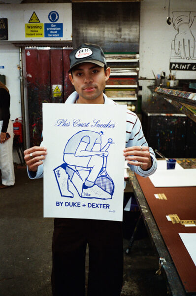 A man in a striped shirt and cap holds a poster with blue line art and text reading "Plus Court Sneaker BY DUKE + DEXTER" in a workshop setting.