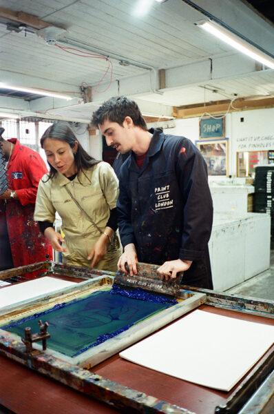 Two people stand at a screen printing station, working together to spread ink across a screen, with blank paper and art supplies visible around them in a well-lit studio.