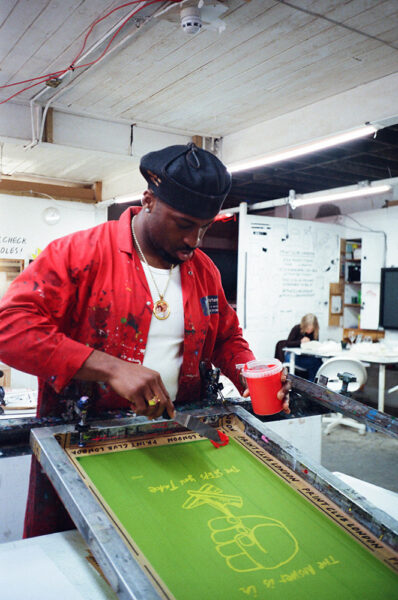 A man in a red coat holds a red cup while screen printing a design with text and a raised fist on a green sheet in an art studio.