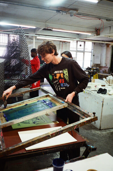 A person in a black graphic shirt is screen printing on paper in an art studio, with another person and printing equipment visible in the background.