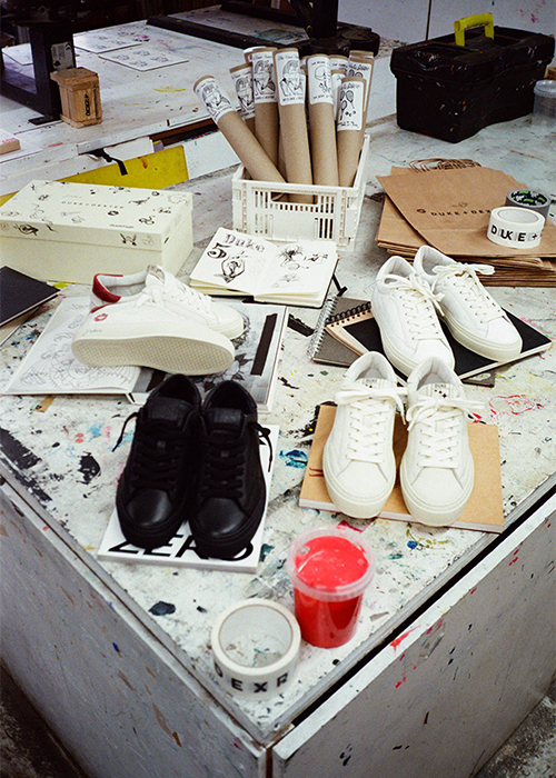 A worktable displays white and black trainers, rolled-up posters, sketches, brown folders, tape, and a cup of red paint in an art studio setting—perfect inspiration for Live-Events-and-Corporate-Workshops seeking creative energy.