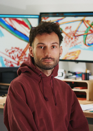 A man in a maroon hoodie, identified as Alex Evans, sits at a desk in front of two monitors displaying colourful abstract art.