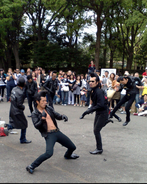 Group of men in black clothing and sunglasses dancing energetically in an outdoor park, with a crowd of people watching in the background.