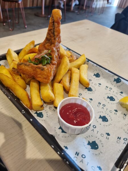 A serving of thick-cut chips and a piece of battered fish with a garnish, accompanied by a small pot of tomato sauce, on a branded tray liner at a restaurant.