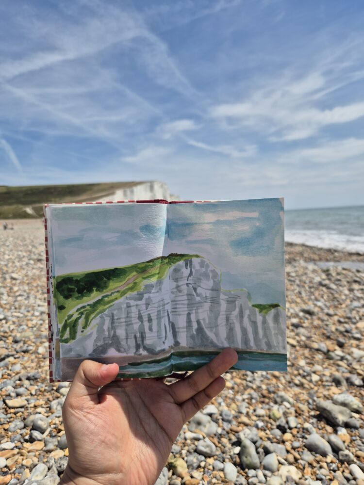 A hand holds an open sketchbook with a painting of white cliffs and sea, matching the real cliffs and shoreline in the background.