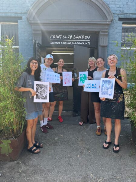 Six people stand outside a print studio in London, each holding a screen print they made, under a sign for Print Club London.