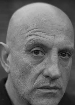 A bald man with a serious expression faces the camera in a black and white close-up portrait.
