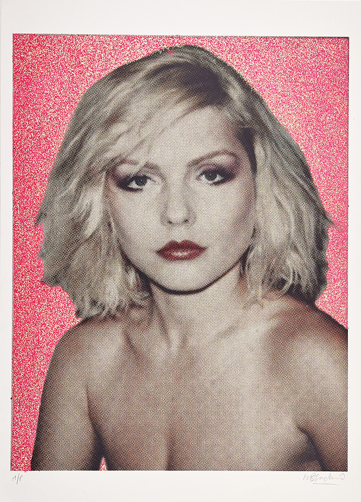 A woman with short blonde hair stands in front of a pink glittery background.